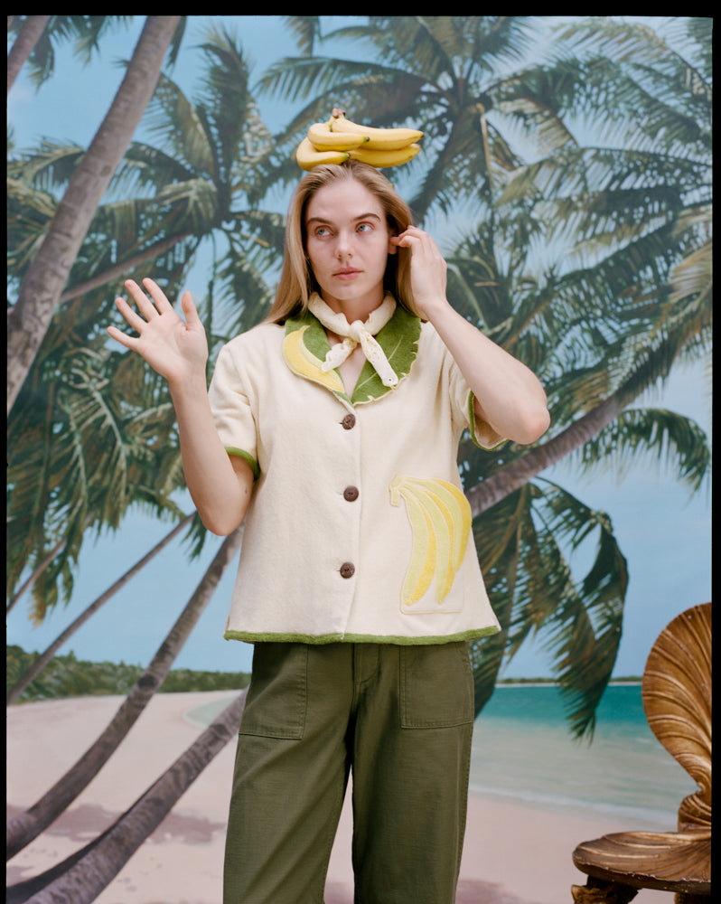 A woman wearing a cream-colored short-sleeve shirt with green trim and a large banana applique on the front, green pants, and a light scarf tied around her neck. She has a bunch of bananas sitting on top of her head. 