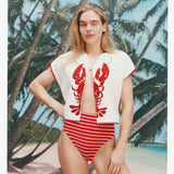 Female model with long blonde hair winking at the camera while wearing the 'Better Half' Hand Towel Top with the top button buttoned and a red white striped bikini bottom, shot against a bacgkround of palm trees, blue skies, and a beach.