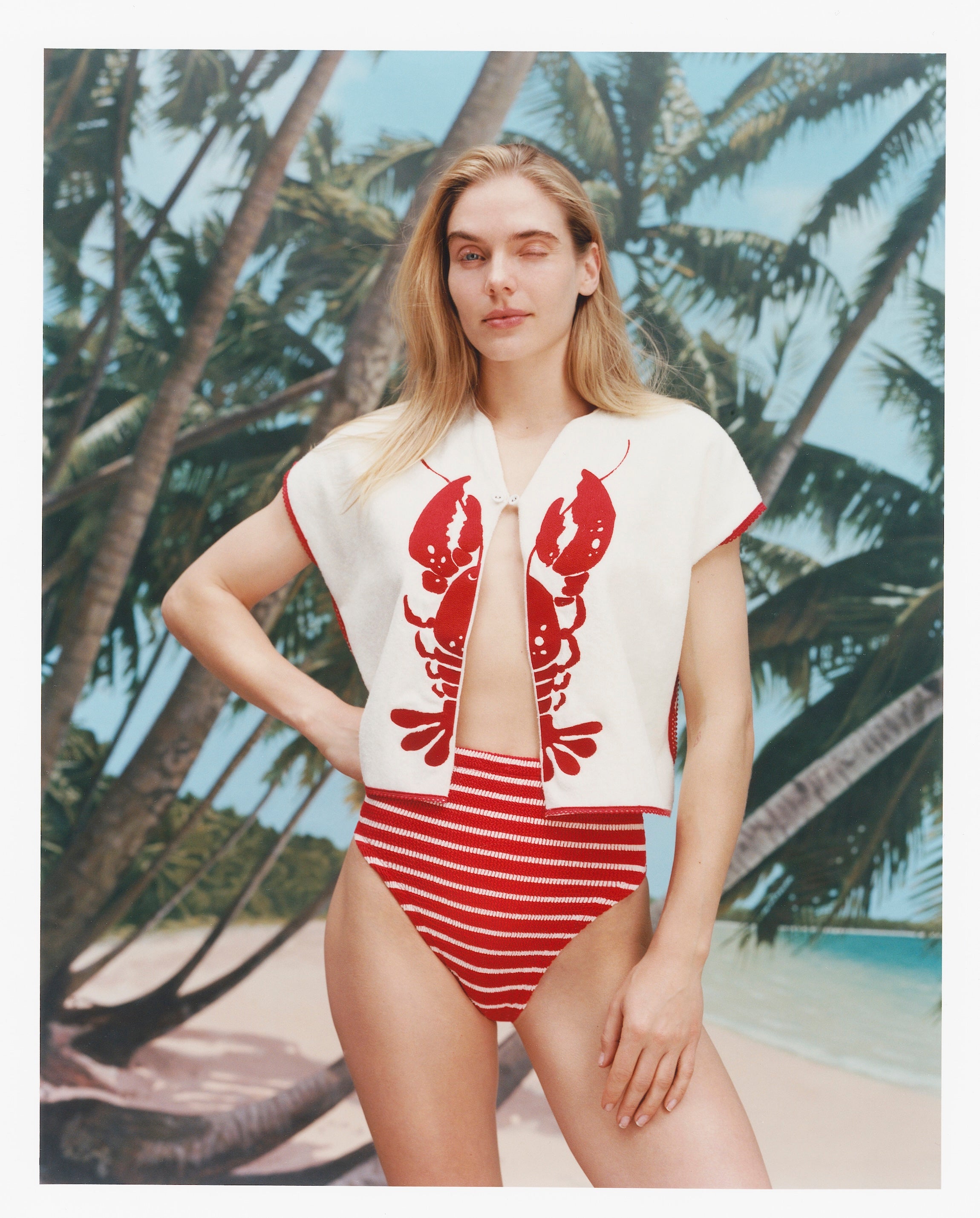 Female model with long blonde hair winking at the camera while wearing the 'Better Half' Hand Towel Top with the top button buttoned and a red white striped bikini bottom, shot against a bacgkround of palm trees, blue skies, and a beach.