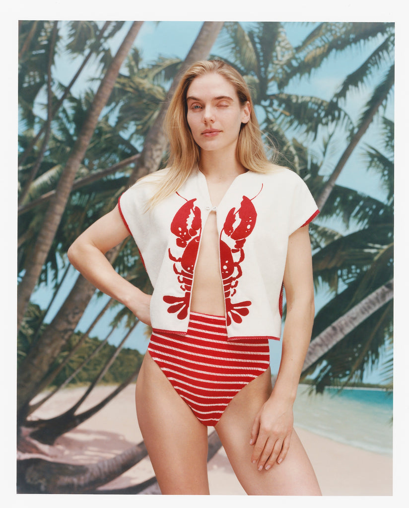 Female model with long blonde hair winking at the camera while wearing the 'Better Half' Hand Towel Top with the top button buttoned and a red white striped bikini bottom, shot against a bacgkround of palm trees, blue skies, and a beach.