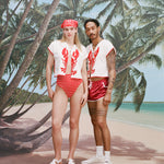 Female model with long blonde hair and male model with dreadlocks posing together both wearing the 'Better Half' Hand Towel Top with the top button buttoned, the female model has a red and white cloth covering her hair, and is wearing a red and white striped bikini bottom, and white sandals, while the male model is wearing a pair of red 'short-shorts' with white trim, white socks, and sneakers, all shot against a background of palm trees, blue skies, and a beach