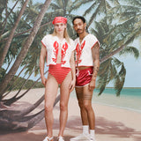Female model with long blonde hair and male model with dreadlocks posing together both wearing the 'Better Half' Hand Towel Top with the top button buttoned, the female model has a red and white cloth covering her hair, and is wearing a red and white striped bikini bottom, and white sandals, while the male model is wearing a pair of red 'short-shorts' with white trim, white socks, and sneakers, all shot against a background of palm trees, blue skies, and a beach