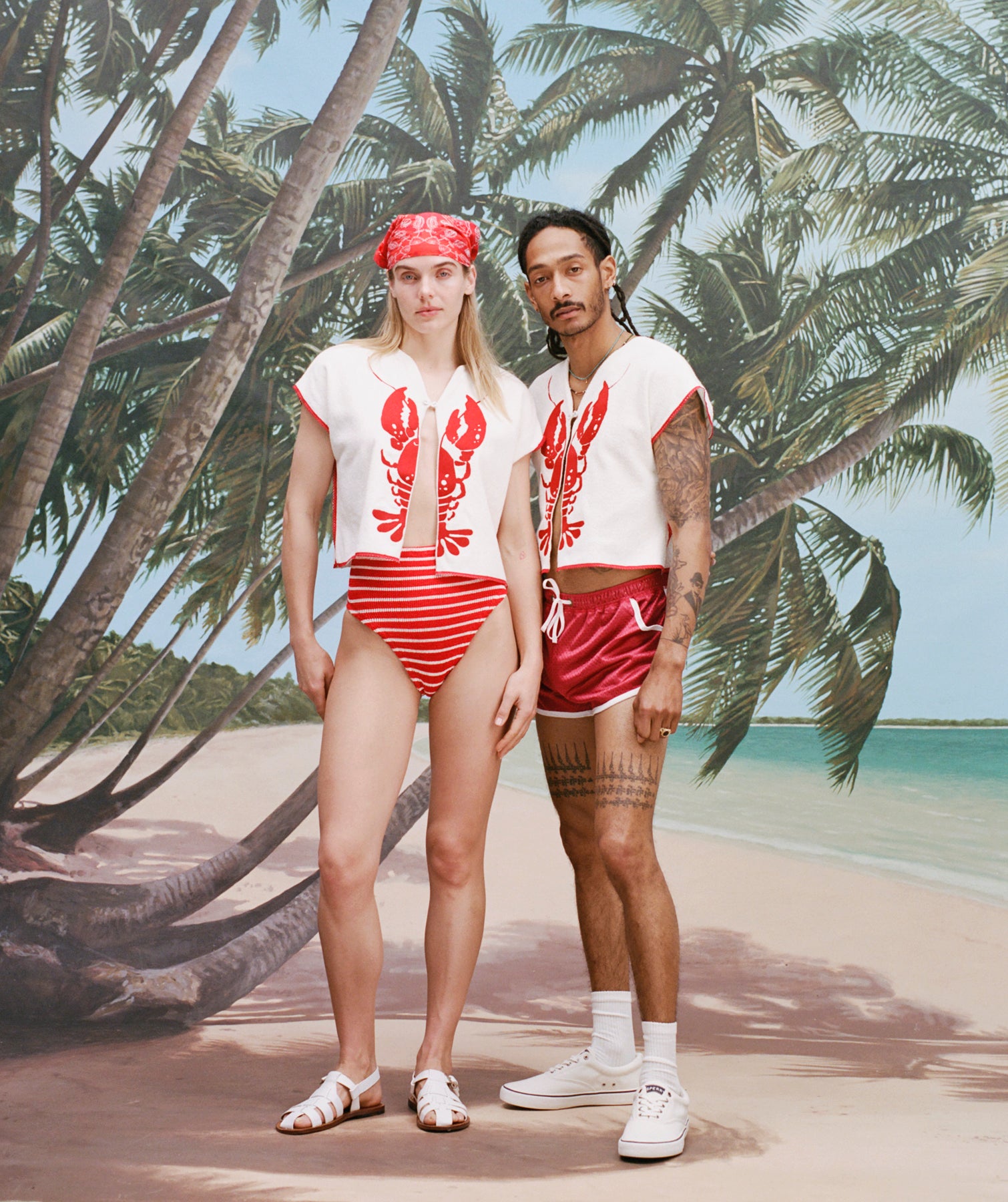 Female model with long blonde hair and male model with dreadlocks posing together both wearing the 'Better Half' Hand Towel Top with the top button buttoned, the female model has a red and white cloth covering her hair, and is wearing a red and white striped bikini bottom, and white sandals, while the male model is wearing a pair of red 'short-shorts' with white trim, white socks, and sneakers, all shot against a background of palm trees, blue skies, and a beach