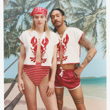 Female model with long blonde hair and male model with dreadlocks pose together with both wearing the 'Better Half' Hand Towel Top with the top button buttoned, the female model is wearing a red and white colored cloth covering her hair, and a red and white striped bikini bottom, while the male model is wearing a pair of red 'short-shorts' with white trim, all shot against a background of palm trees, blue skies, and a beach.