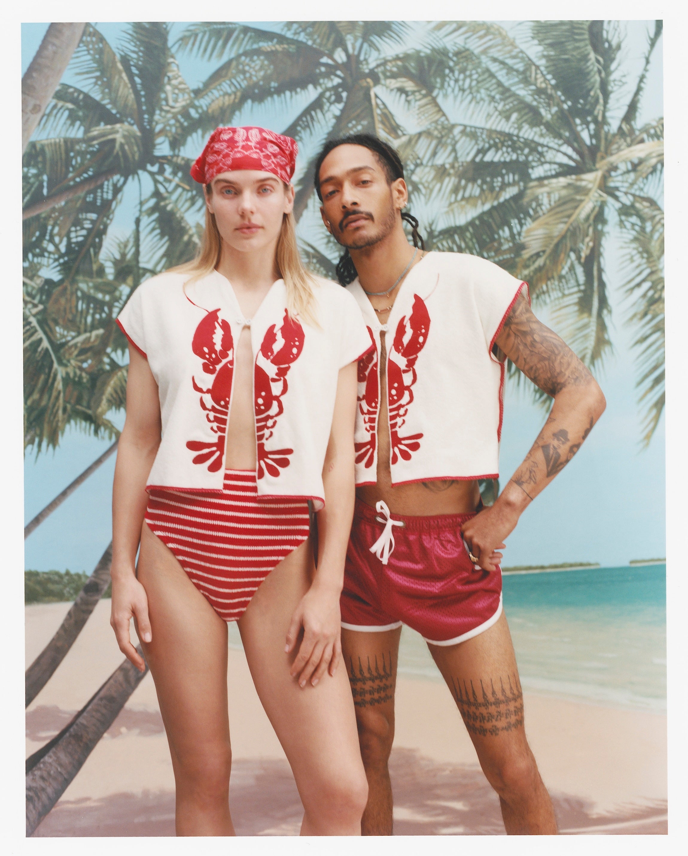 Female model with long blonde hair and male model with dreadlocks pose together with both wearing the 'Better Half' Hand Towel Top with the top button buttoned, the female model is wearing a red and white colored cloth covering her hair, and a red and white striped bikini bottom, while the male model is wearing a pair of red 'short-shorts' with white trim, all shot against a background of palm trees, blue skies, and a beach.