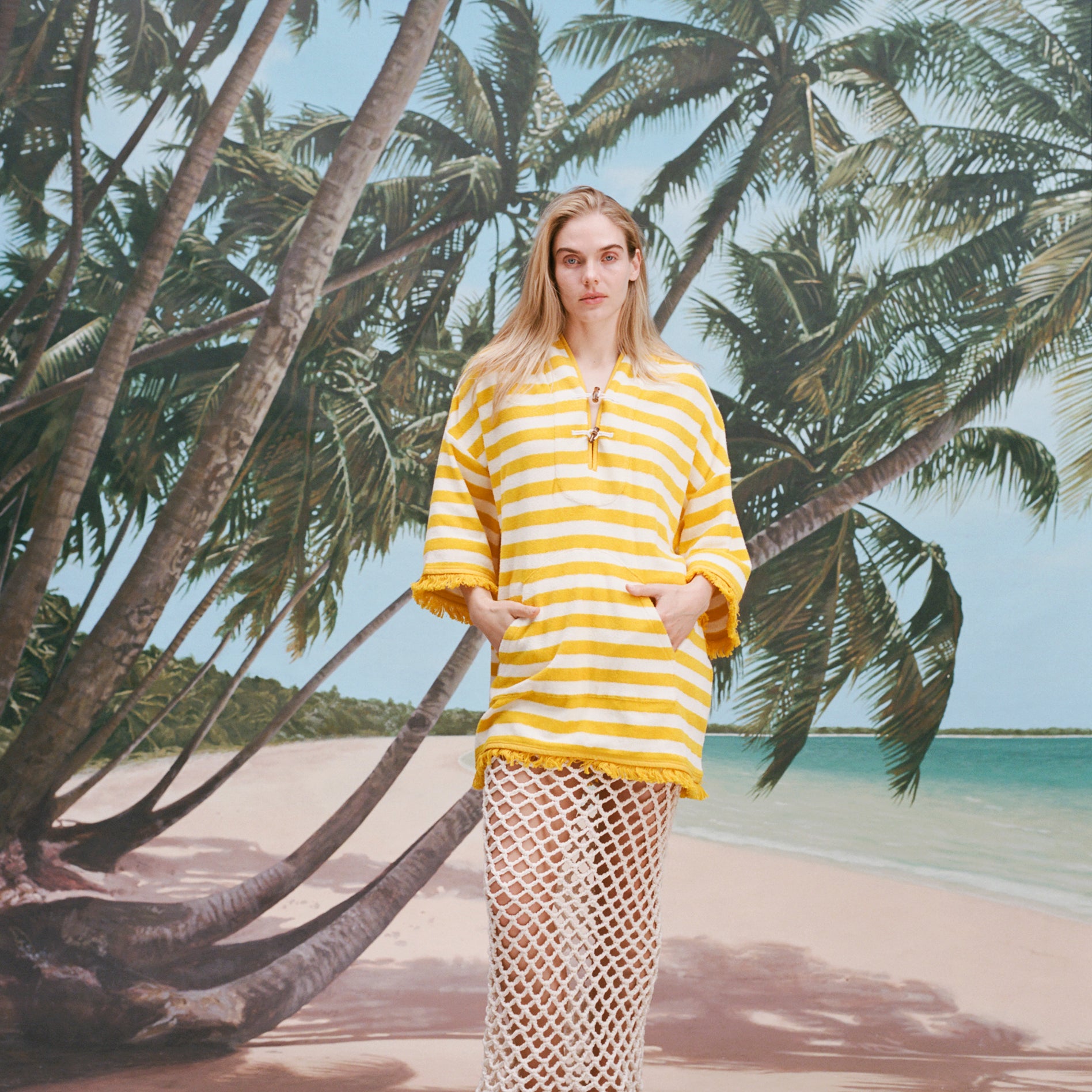 Female model with long blonde hair wearing the 'Saffron Stripe' Beach Towel top, a white skirt with netting, white sandals, shot on a background of a beach, palm fronds, and blue skies.