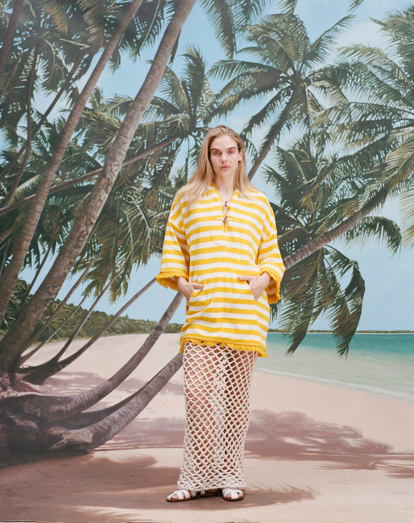 Female model with long blonde hair wearing the 'Saffron Stripe' Beach Towel top, a white skirt with netting, white sandals, shot on a background of a beach, palm fronds, and blue skies.