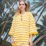 Female model with long blonde hair wearing the 'Saffron Stripe' beach towel top, shot against a background of palm fronds and blue skies
