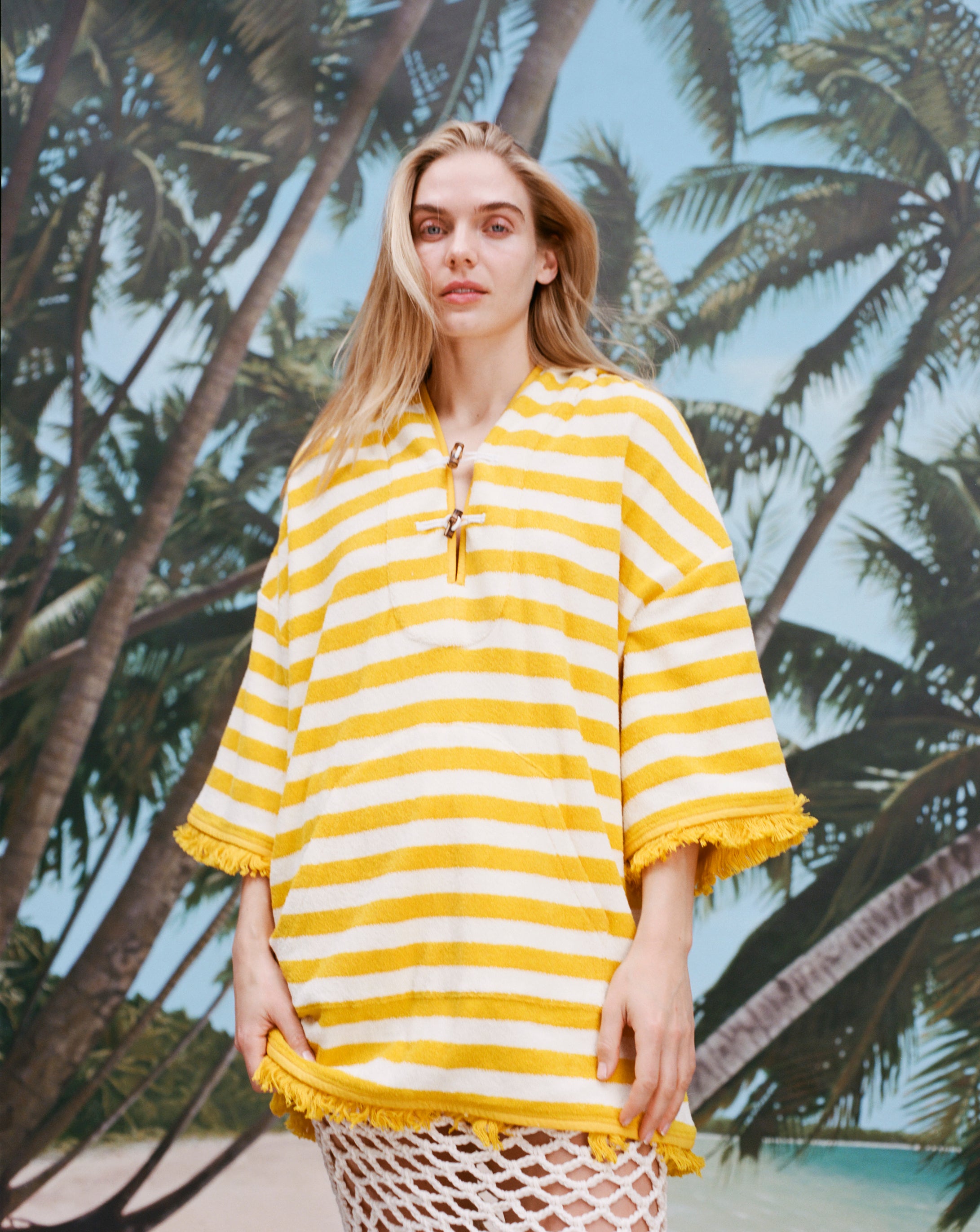 Female model with long blonde hair wearing the 'Saffron Stripe' beach towel top, shot against a background of palm fronds and blue skies