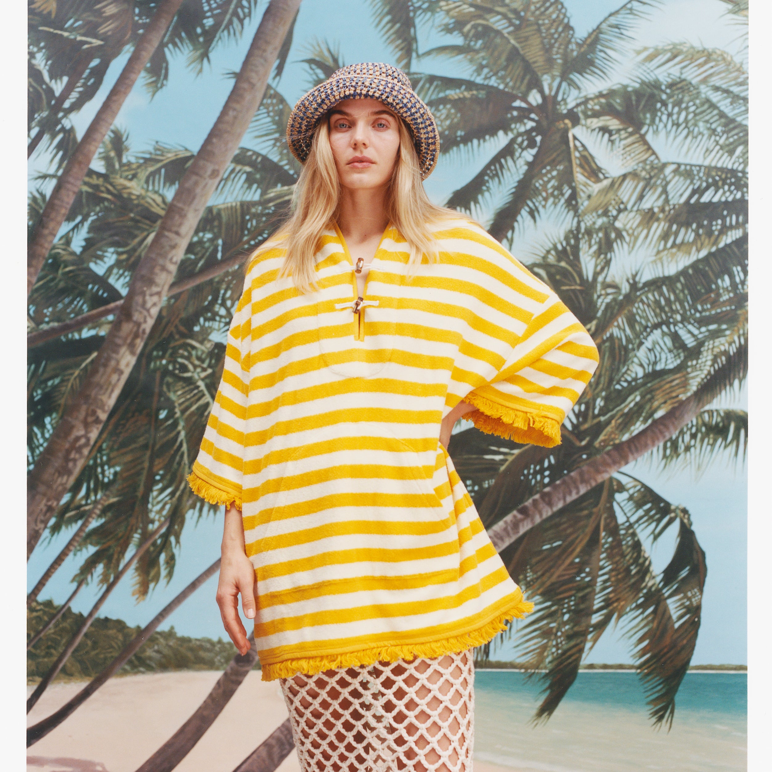 Female model with long blonde hair wearing the 'saffron stripe' beach towel top with a skirt with wide netting holes, and a sun hat, shot on a background of blue skies, a beach, and palm fronds.