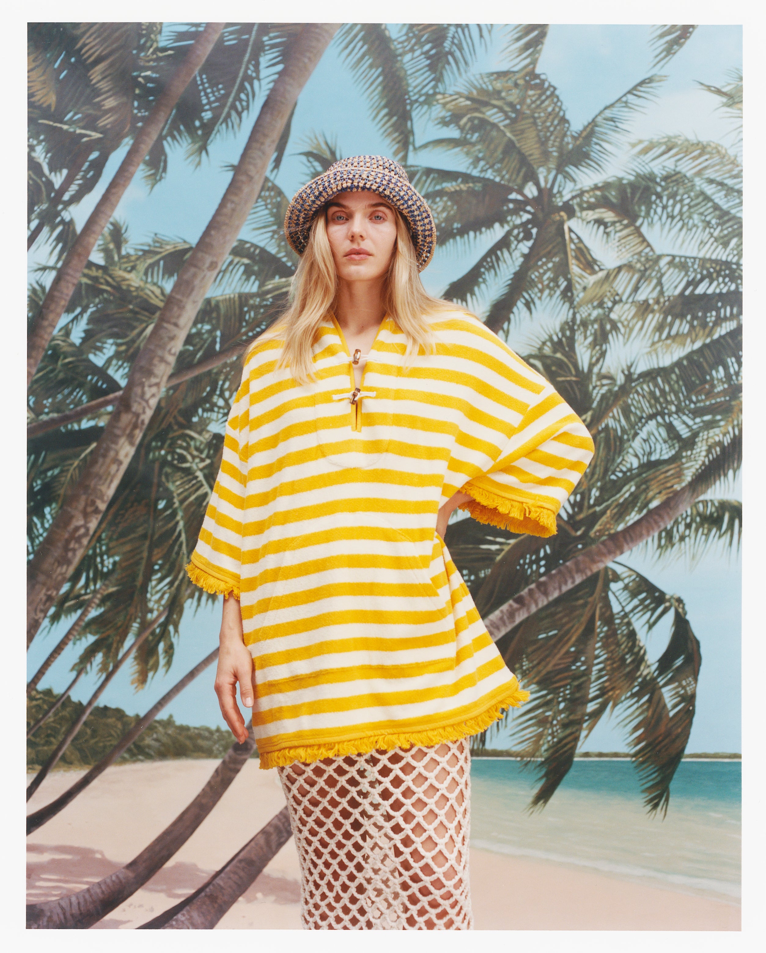 Female model with long blonde hair wearing the 'saffron stripe' beach towel top with a skirt with wide netting holes, and a sun hat, shot on a background of blue skies, a beach, and palm fronds.