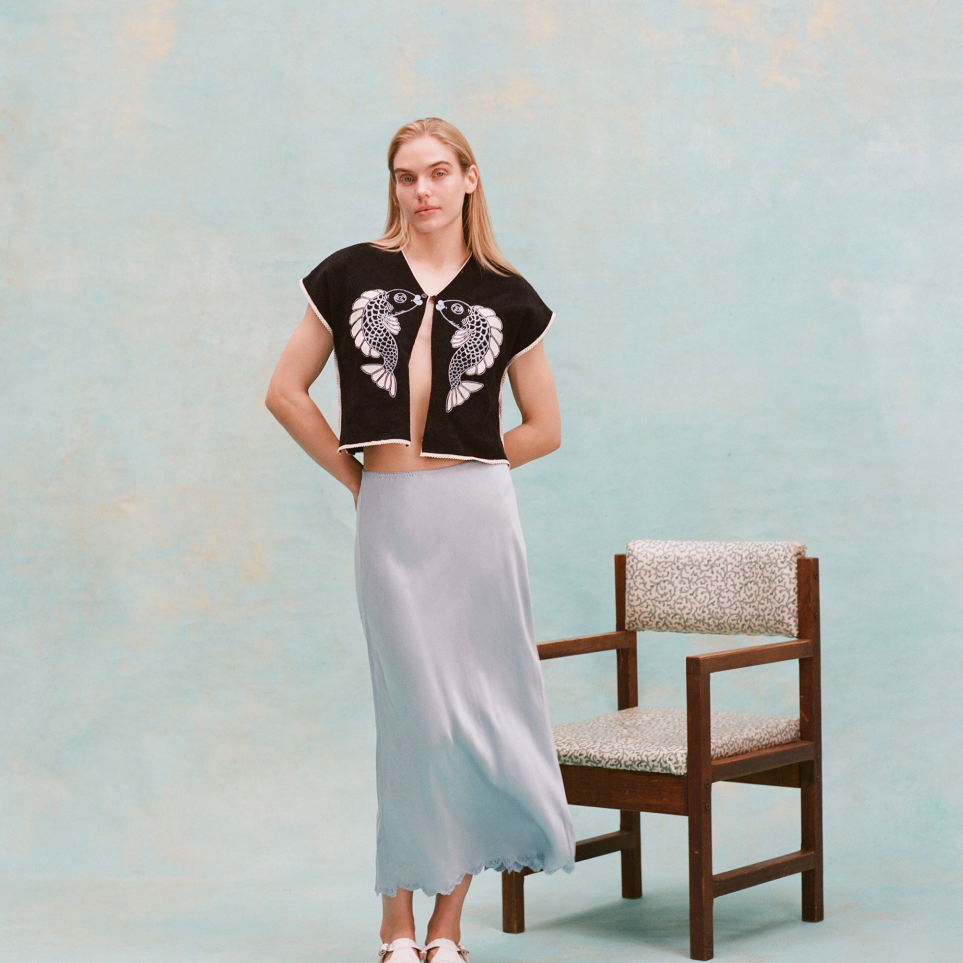 Female model with long blonde hair wearing the 'fish smooch' hand towel top with a light blue silk dress and white sandals, shot on a light blue textured background.