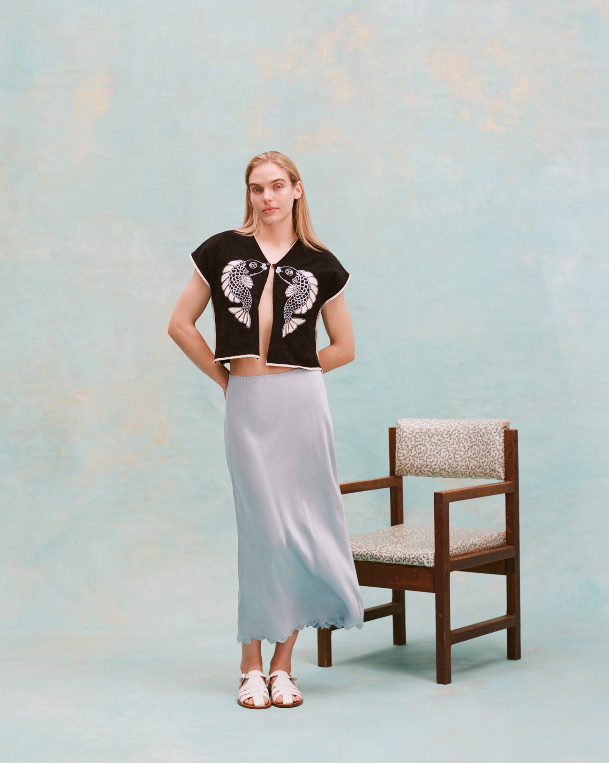 Female model with long blonde hair wearing the 'fish smooch' hand towel top with a light blue silk dress and white sandals, shot on a light blue textured background.