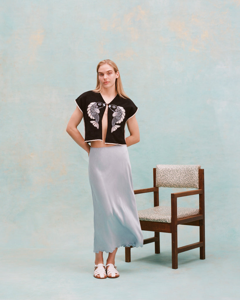 Female model with long blonde hair wearing the 'fish smooch' hand towel top with a light blue silk dress and white sandals, shot on a light blue textured background.