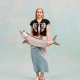 Female model with long blonde hair wearing the 'fish smooch' hand towel top and a light blue silk dress with white sandals, holding a plaster fish, shot on a light blue textured background.
