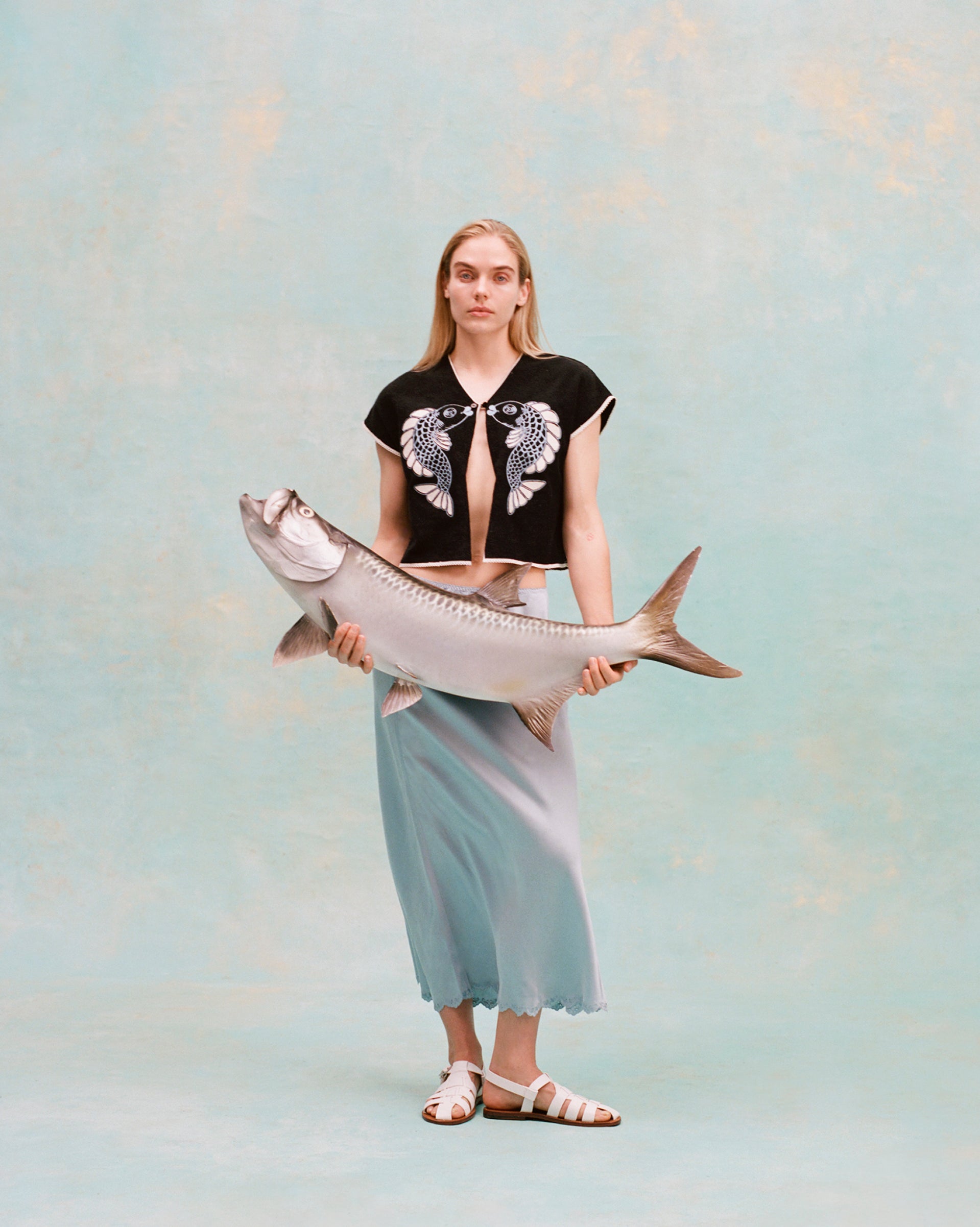 Female model with long blonde hair wearing the 'fish smooch' hand towel top and a light blue silk dress with white sandals, holding a plaster fish, shot on a light blue textured background.