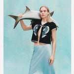 Female model with long blonde hair wearing the 'fish smooch' hand towel top while wearing a light blue silk dress, holding a plaster fish over her shoulder, shot on a textured light blue background.
