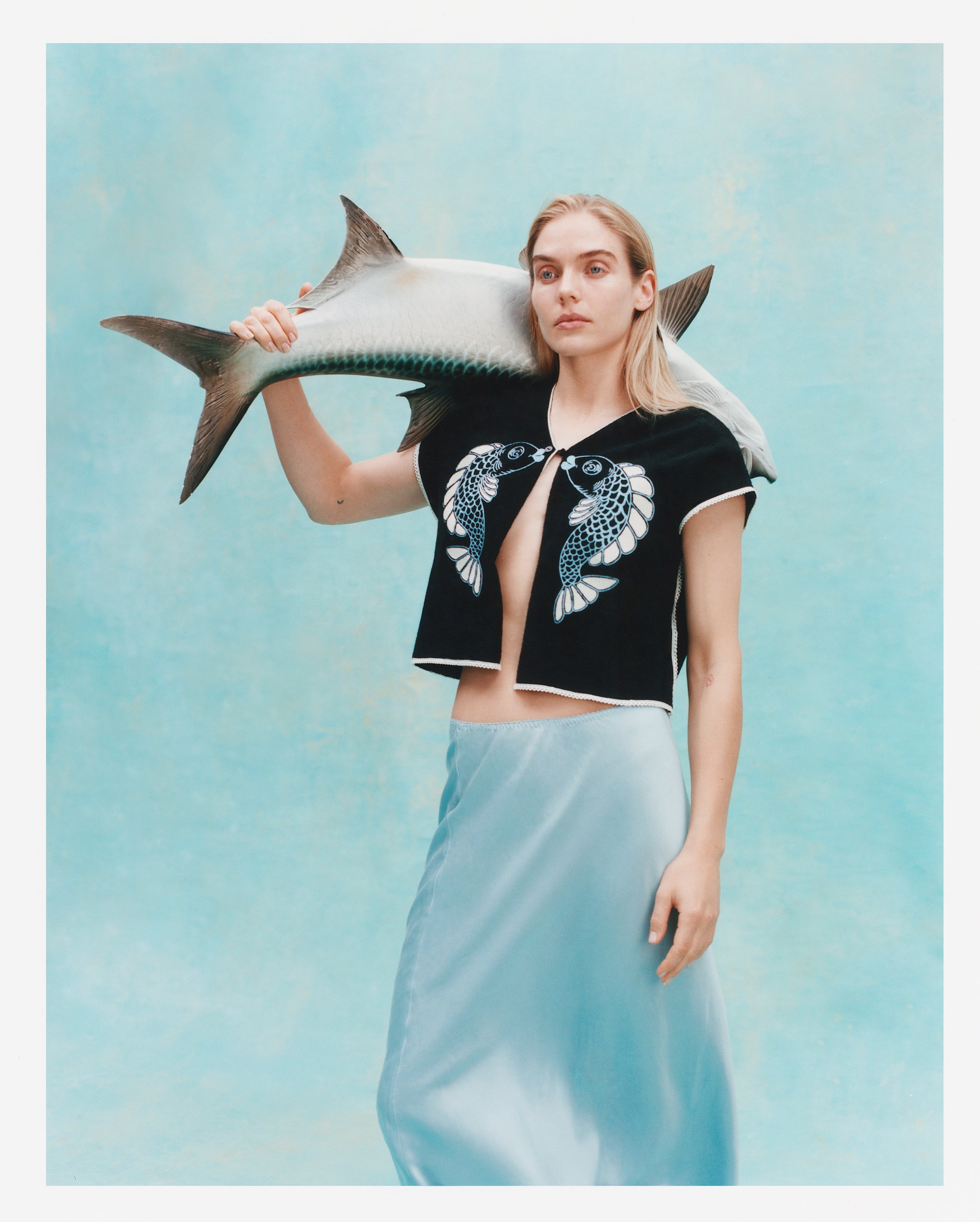 Female model with long blonde hair wearing the 'fish smooch' hand towel top while wearing a light blue silk dress, holding a plaster fish over her shoulder, shot on a textured light blue background.