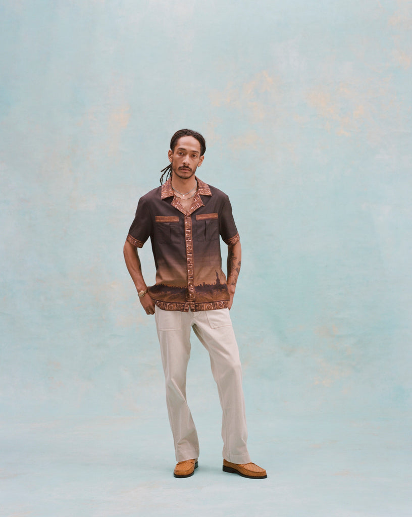 Young male model wearing 'The Sopranos' Turnpike Batik with both of his hands in his pockets, one leg slightly bent forward, in khakis, and brown moccasins, looking towards the camera, shot on a light baby blue background.