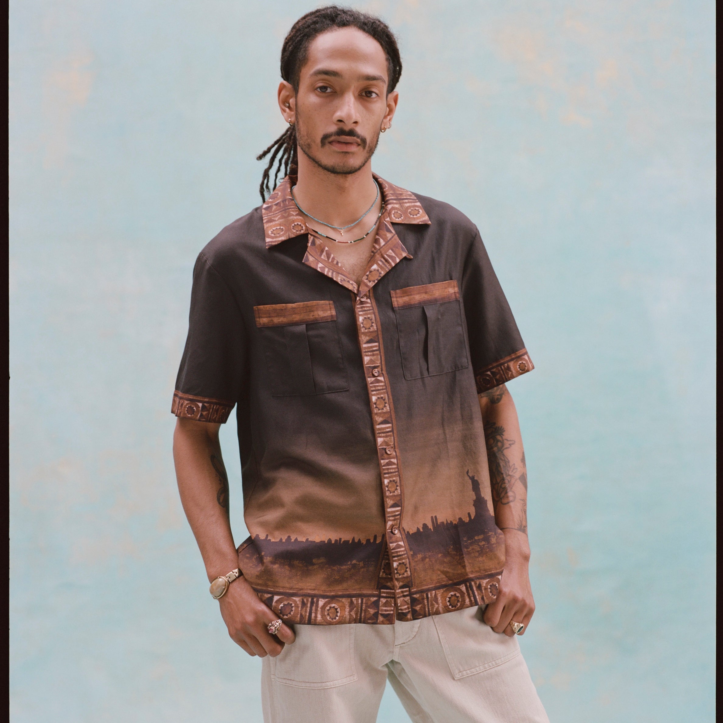 Young male model with both hands in his pockets, wearing khaki pants and 'The Sopranos' Turnpike Batik, on a light baby blue background.