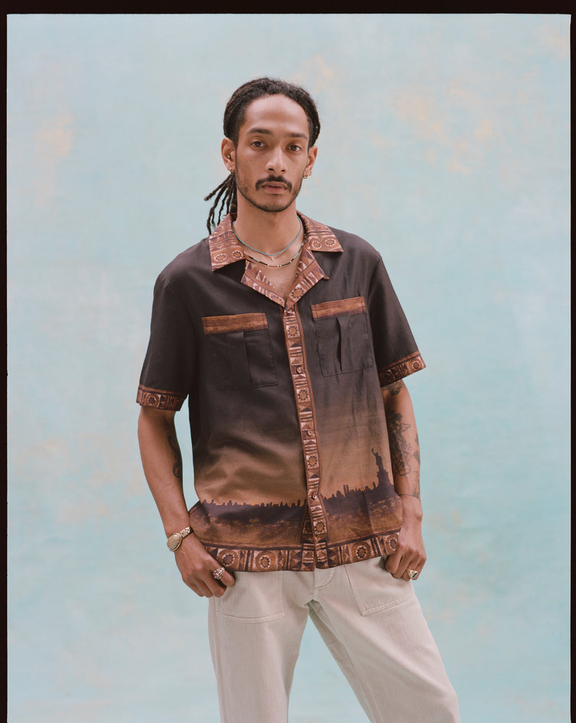 Young male model with both hands in his pockets, wearing khaki pants and 'The Sopranos' Turnpike Batik, on a light baby blue background.