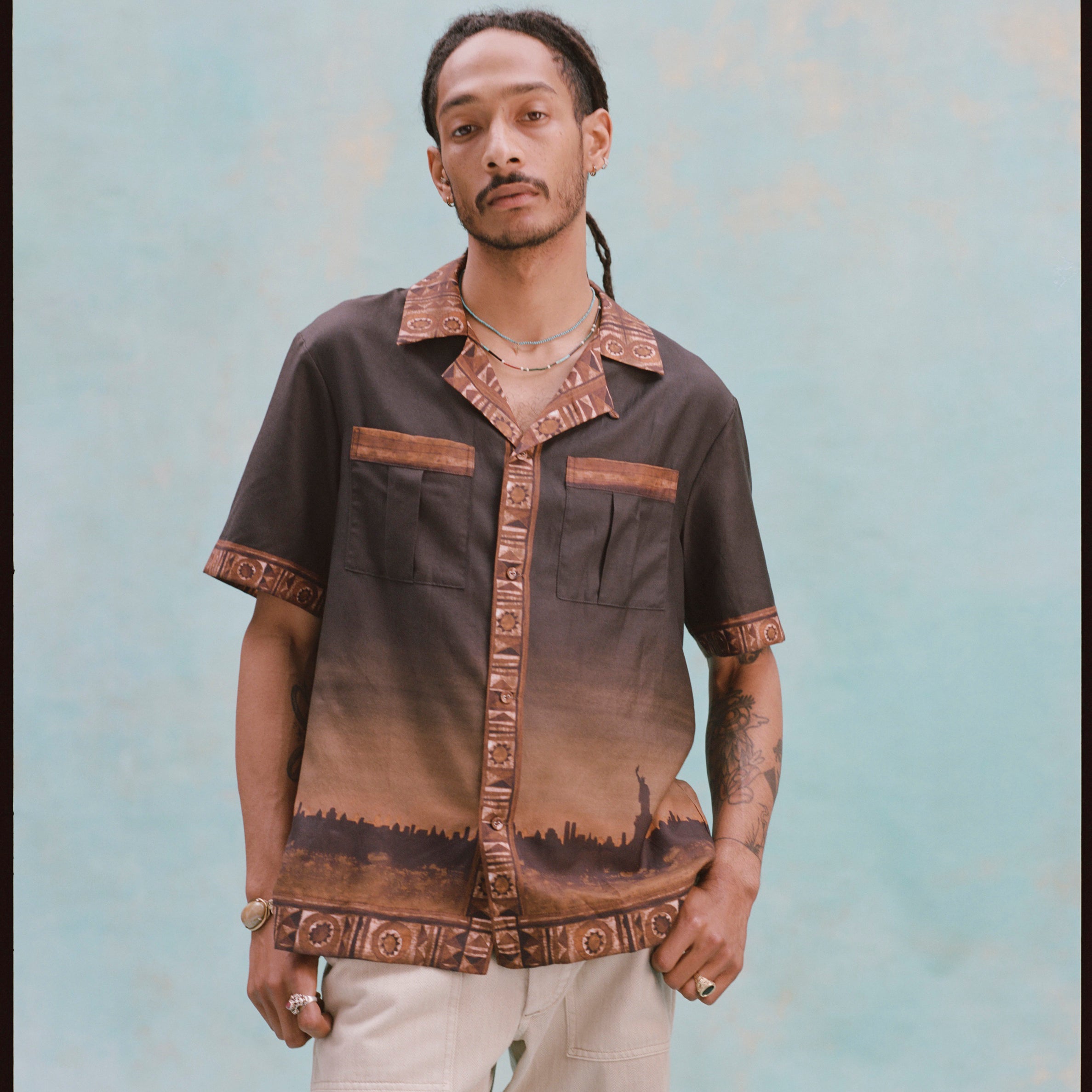 Young male model wearing 'The Sopranos' Turnpike Batik with both hands in his pockets, alongside khaki pants, shot on a very light baby blue background.