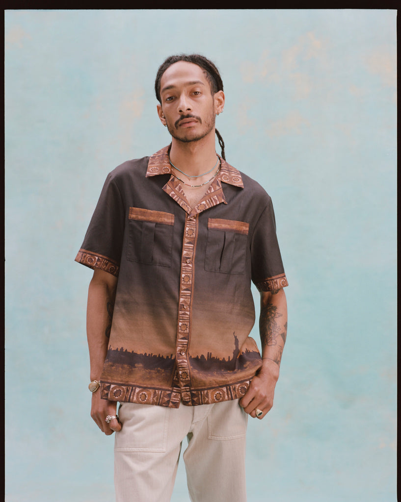 Young male model wearing 'The Sopranos' Turnpike Batik with both hands in his pockets, alongside khaki pants, shot on a very light baby blue background.
