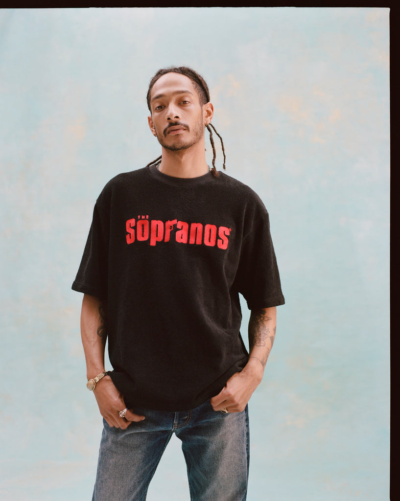 Male model with dreadlocks is standing with his hands in his pockets, wearing a black "The Sopranos" Souvenir Terry Tee and jeans, against a light blue textured background.