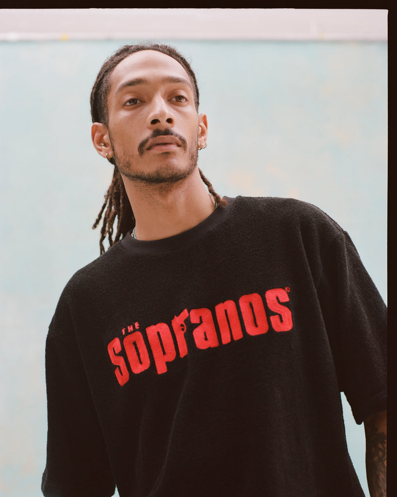 Close-up shot of a young male model with dreadlocks wearing 'The Sopranos' Souvenir Terry Tee that features a red chain-stitched 'The Sopranos' logo across chest on a black terry cloth tee.