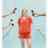 Woman wearing a matching orange terry cloth cabana shirt and shorts set with white embroidered lobsters on the collar and coiled telephone cord details. Several hands surround her holding vintage telephone receivers.