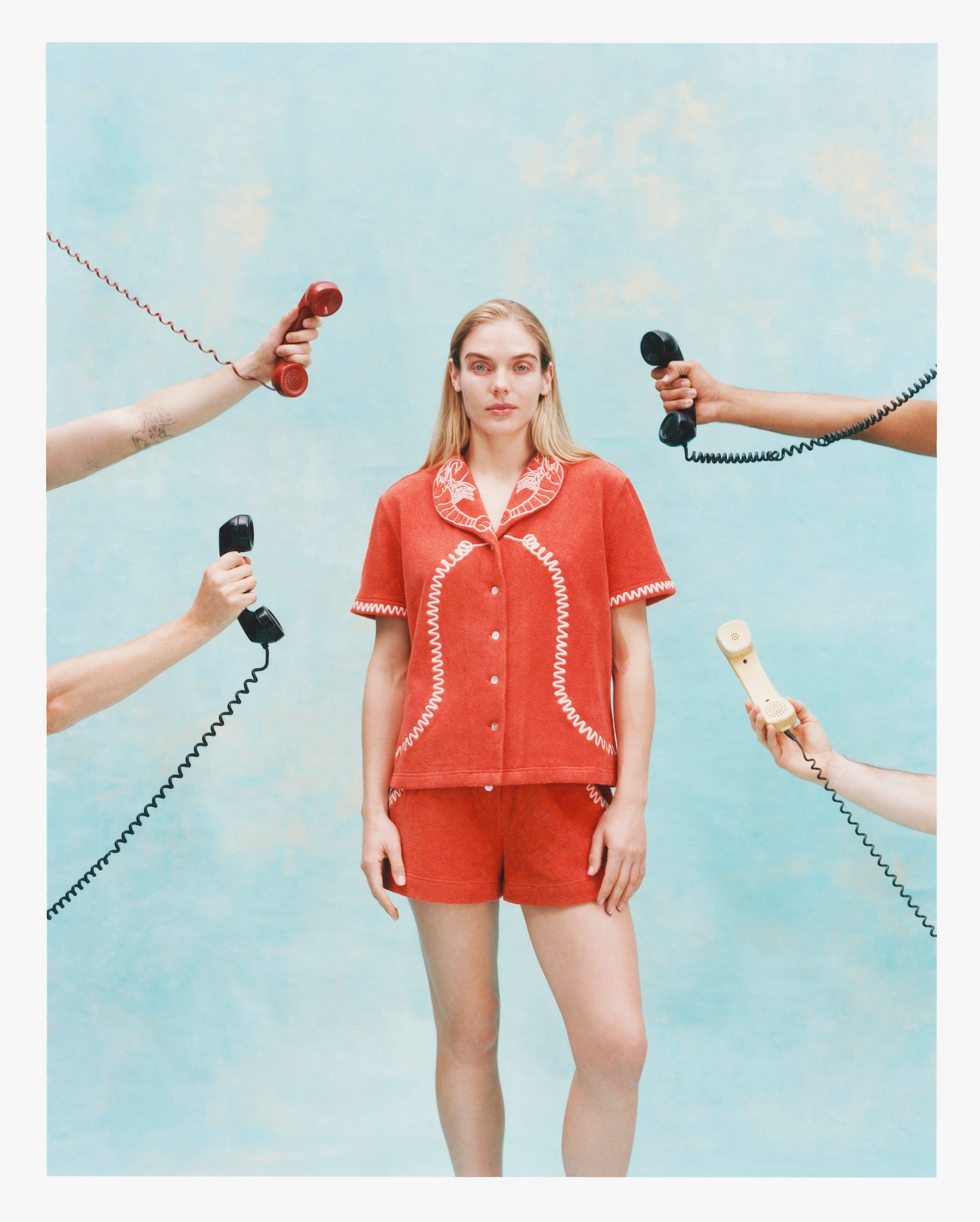 Woman wearing a matching orange terry cloth cabana shirt and shorts set with white embroidered lobsters on the collar and coiled telephone cord details. Several hands surround her holding vintage telephone receivers.