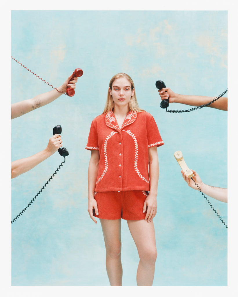 Woman wearing a matching orange terry cloth cabana shirt and shorts set with white embroidered lobsters on the collar and coiled telephone cord details. Several hands surround her holding vintage telephone receivers.