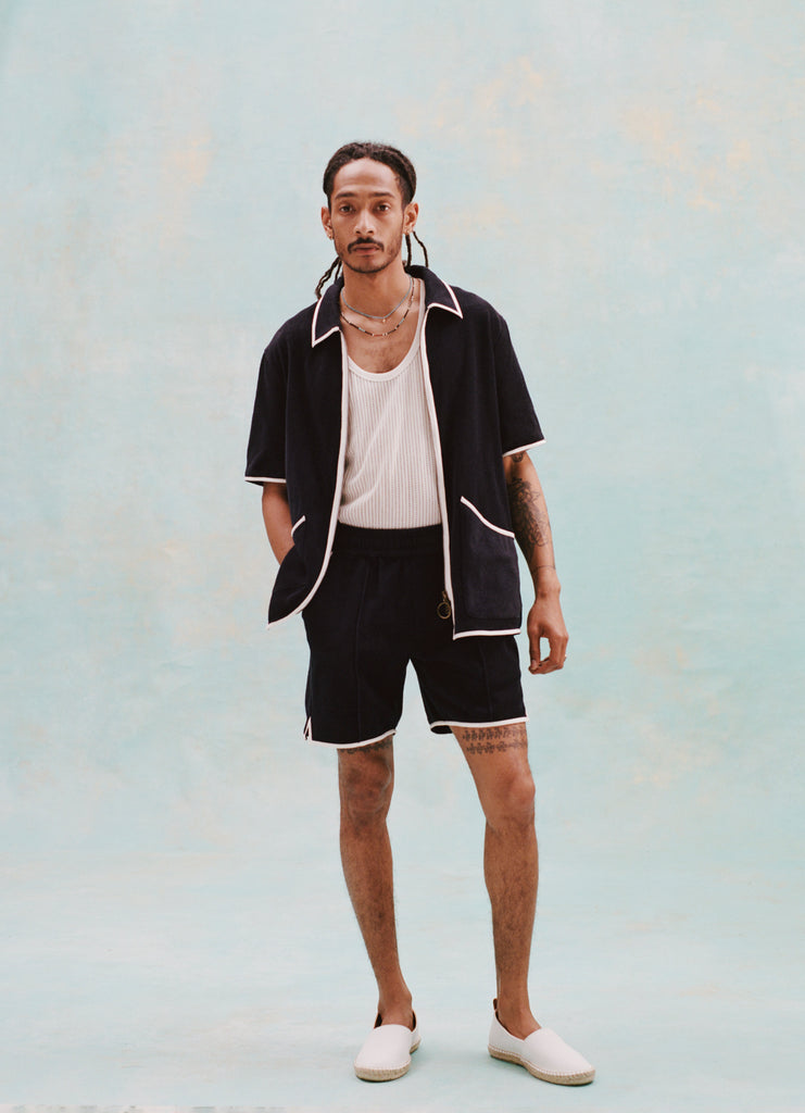 Male model with dreadlocks wearing the 'Standard Issue' Cabana (Midnight Blue) fully unzipped with a white tank top beneath it, matching 'standard issue' shorts in midnight blue, and white slippers, all shot on a white background.