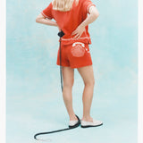 Back view of a woman wearing a matching orange terry cloth shirt and shorts set with white embroidered details, including a telephone and seashell motif on the shorts. 