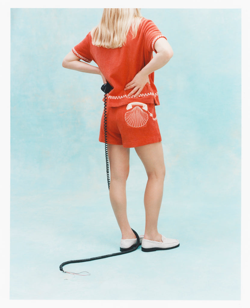 Back view of a woman wearing a matching orange terry cloth shirt and shorts set with white embroidered details, including a telephone and seashell motif on the shorts. 