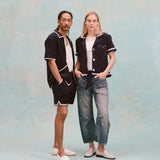 Male model with dreadlocks and female model with long blonde hair posing together, the male model is wearing the 'Standard Issue' Cabana (midnight blue) with a white tank top underneath and matching 'standard issue' shorts in midnight blue with white slippers while the female model is wearing the 'Rick Rack' Cabana ~ Crop Cut (Midnight Blue) unbuttoned, but tied at the waist, with a white shirt underneath and white paint splattered jeans with white loafers, all shot on a white background.