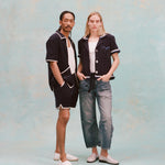Male model with dreadlocks and female model with long blonde hair posing together, the male model is wearing the 'Standard Issue' Cabana (midnight blue) with a white tank top underneath and matching 'standard issue' shorts in midnight blue with white slippers while the female model is wearing the 'Rick Rack' Cabana ~ Crop Cut (Midnight Blue) unbuttoned, but tied at the waist, with a white shirt underneath and white paint splattered jeans with white loafers, all shot on a white background.