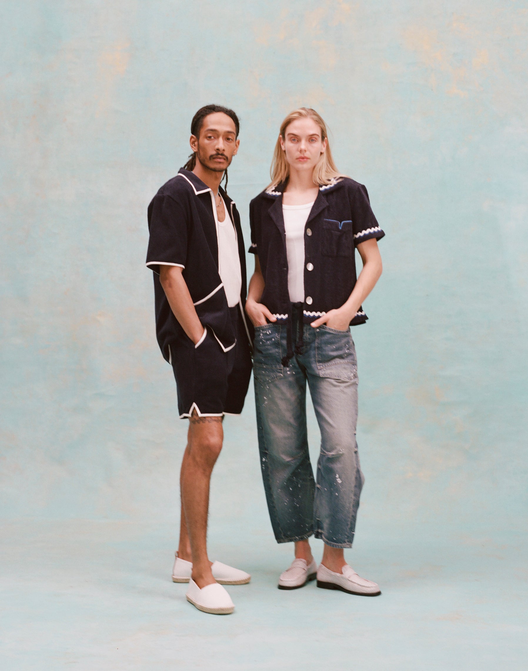Male model with dreadlocks and female model with long blonde hair posing together, the male model is wearing the 'Standard Issue' Cabana (midnight blue) with a white tank top underneath and matching 'standard issue' shorts in midnight blue with white slippers while the female model is wearing the 'Rick Rack' Cabana ~ Crop Cut (Midnight Blue) unbuttoned, but tied at the waist, with a white shirt underneath and white paint splattered jeans with white loafers, all shot on a white background.