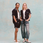 Male model with dreadlocks and female model with long blonde hair posing together, the male model is wearing the 'Standard Issue' Cabana (midnight blue) with a white tank top underneath and matching 'standard issue' shorts in midnight blue with white slippers while the female model is wearing the 'Rick Rack' Cabana ~ Crop Cut (Midnight Blue) unbuttoned, but tied at the waist, with a white shirt underneath and white paint splattered jeans with white loafers, all shot on a white background.