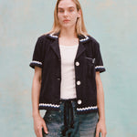 Female model with long blonde hair wearing the 'Rick Rack' Cabana ~ Crop Cut (Midnight blue) unbuttoned with a white shirt, white paint-speckled jeans, and a navy rope belt, shot on textured light blue background.