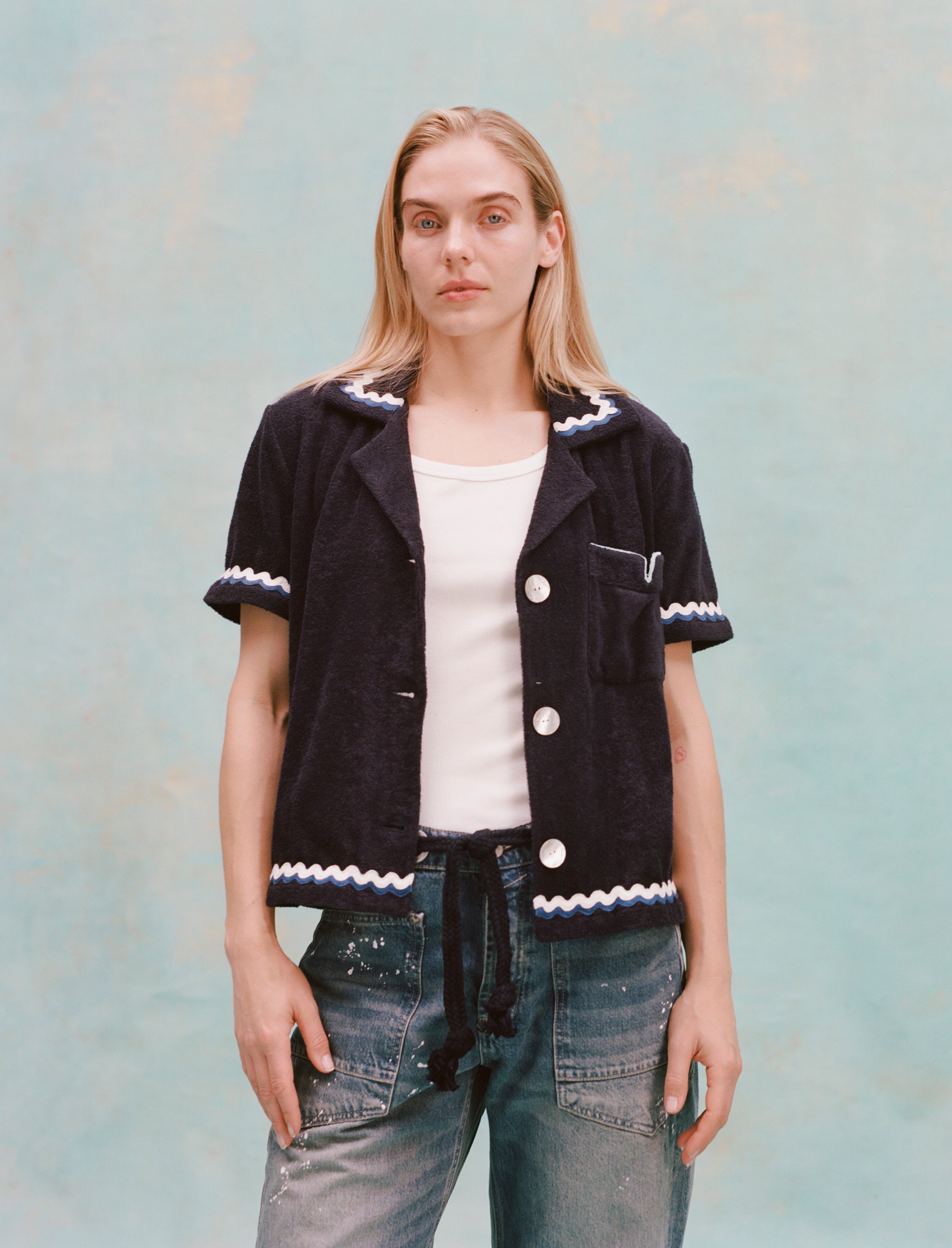 Female model with long blonde hair wearing the 'Rick Rack' Cabana ~ Crop Cut (Midnight blue) unbuttoned with a white shirt, white paint-speckled jeans, and a navy rope belt, shot on textured light blue background.