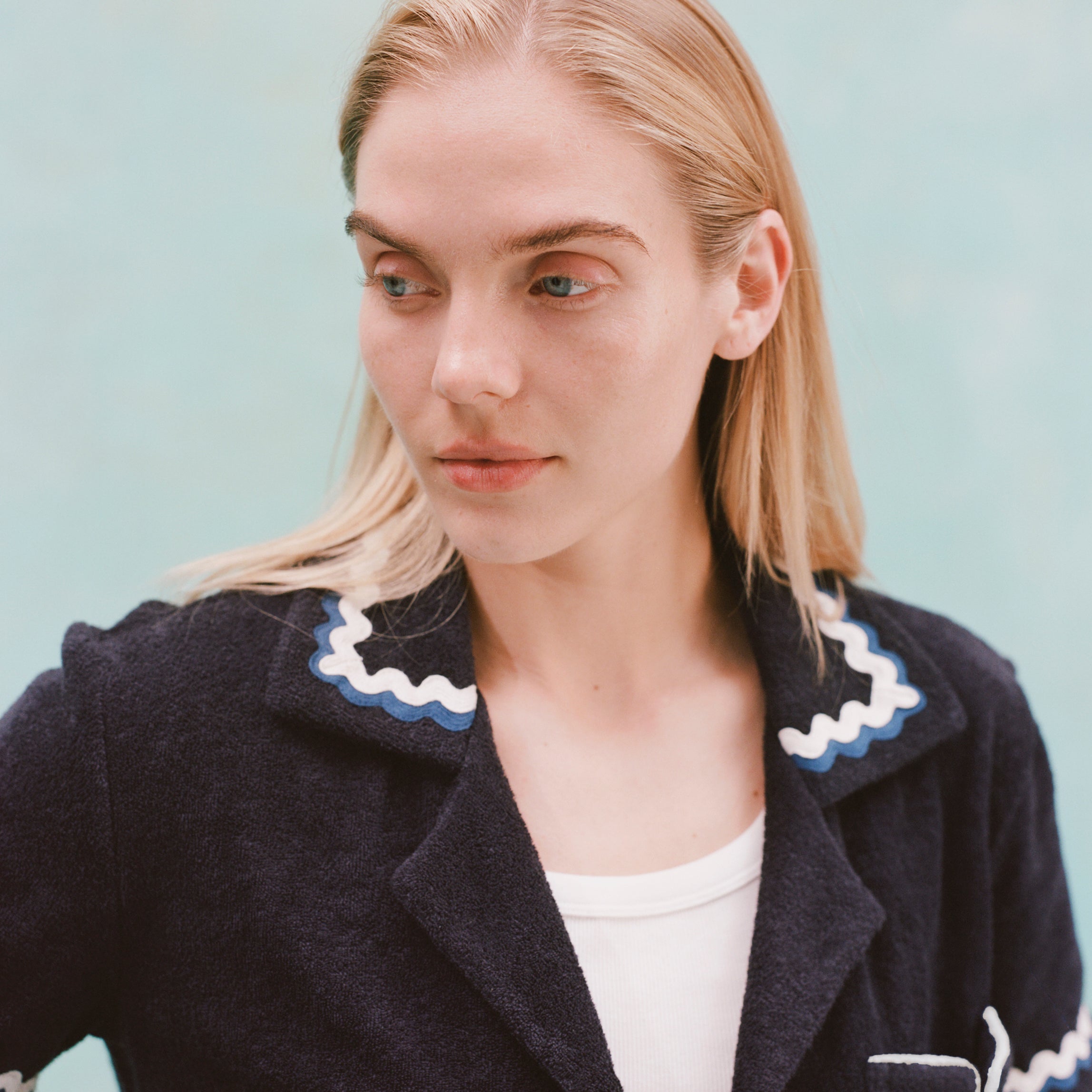 Female model with long blonde hair wearing the 'Rick Rack' Cabana ~ Crop Cut (Midnight Blue) unbuttoned with a white shirt, shot on a textured light blue background.