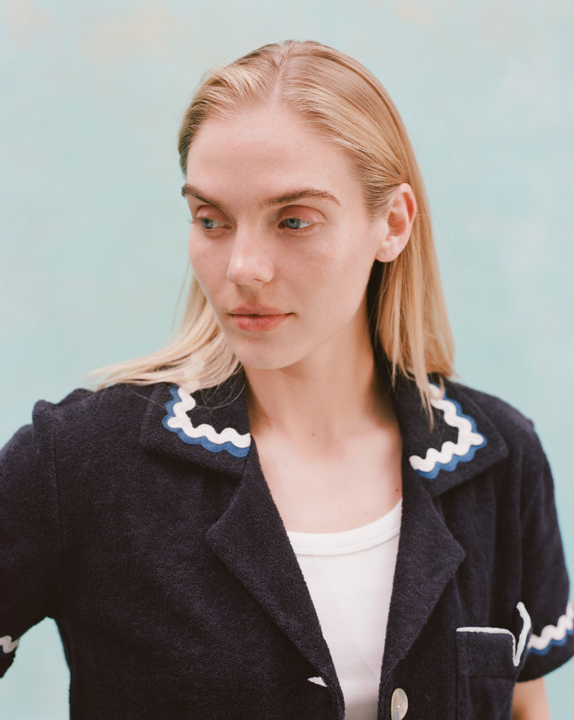 Female model with long blonde hair wearing the 'Rick Rack' Cabana ~ Crop Cut (Midnight Blue) unbuttoned with a white shirt, shot on a textured light blue background.