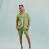 Male model with dreadlocks wearing the 'Standard Issue' Cabana (green) half-zipped with a white, mesh tank top underneath it, and matching 'Standard Issue' green shorts with white slippers, all shot on a light blue background.