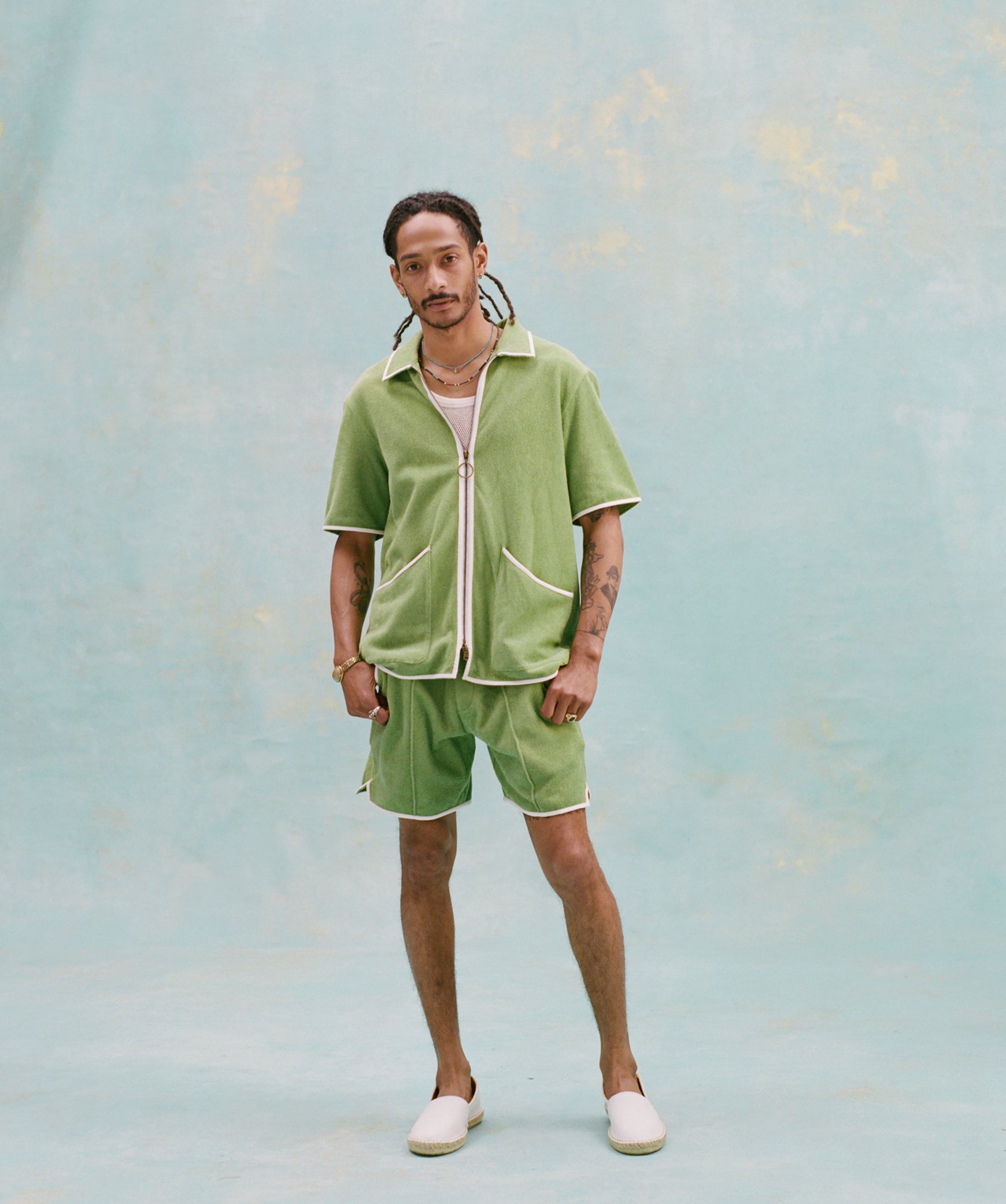 Male model with dreadlocks wearing the 'Standard Issue' Cabana (green) half-zipped with a white, mesh tank top underneath it, and matching 'Standard Issue' green shorts with white slippers, all shot on a light blue background.