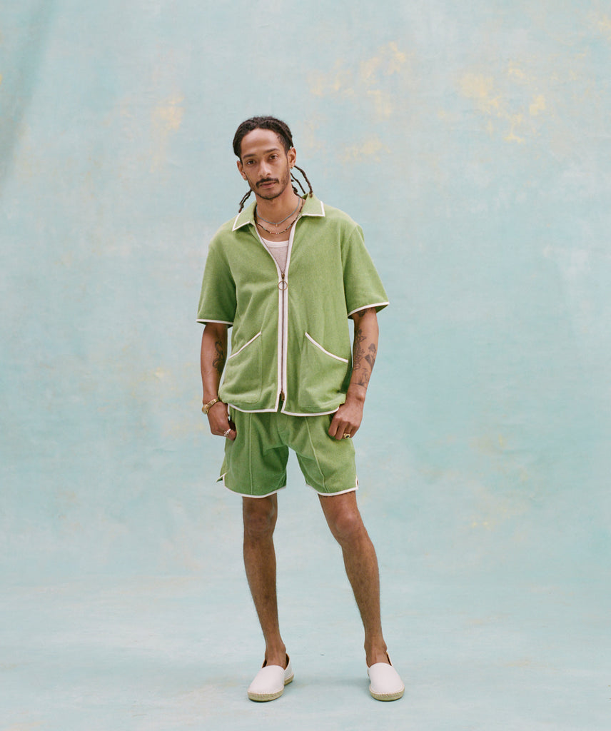 Male model with dreadlocks wearing the 'Standard Issue' Cabana (green) half-zipped with a white, mesh tank top underneath it, and matching 'Standard Issue' green shorts with white slippers, all shot on a light blue background.