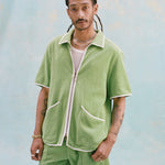 Male model with dreadlocks wearing the 'Standard Issue' Cabana (green) half-zipped with a mesh white tank top underneath and matching 'Standard Issue' green shorts, against a light blue background.