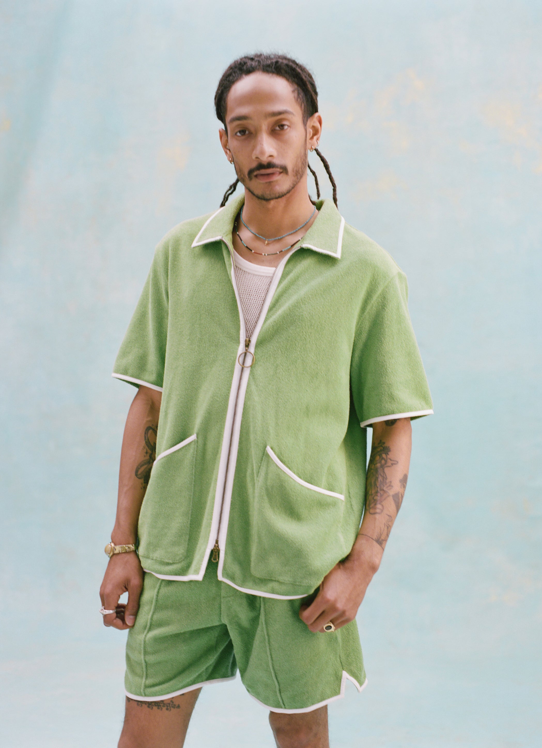 Male model with dreadlocks wearing the 'Standard Issue' Cabana (green) half-zipped with a mesh white tank top underneath and matching 'Standard Issue' green shorts, against a light blue background.