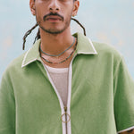 Male model with dreadlocks wearing the 'Standard Issue' Cabana (green) half-zipped with a mesh, white tank top beneath it, and two beaded multicolored necklaces around his neck, shot on a light blue background.