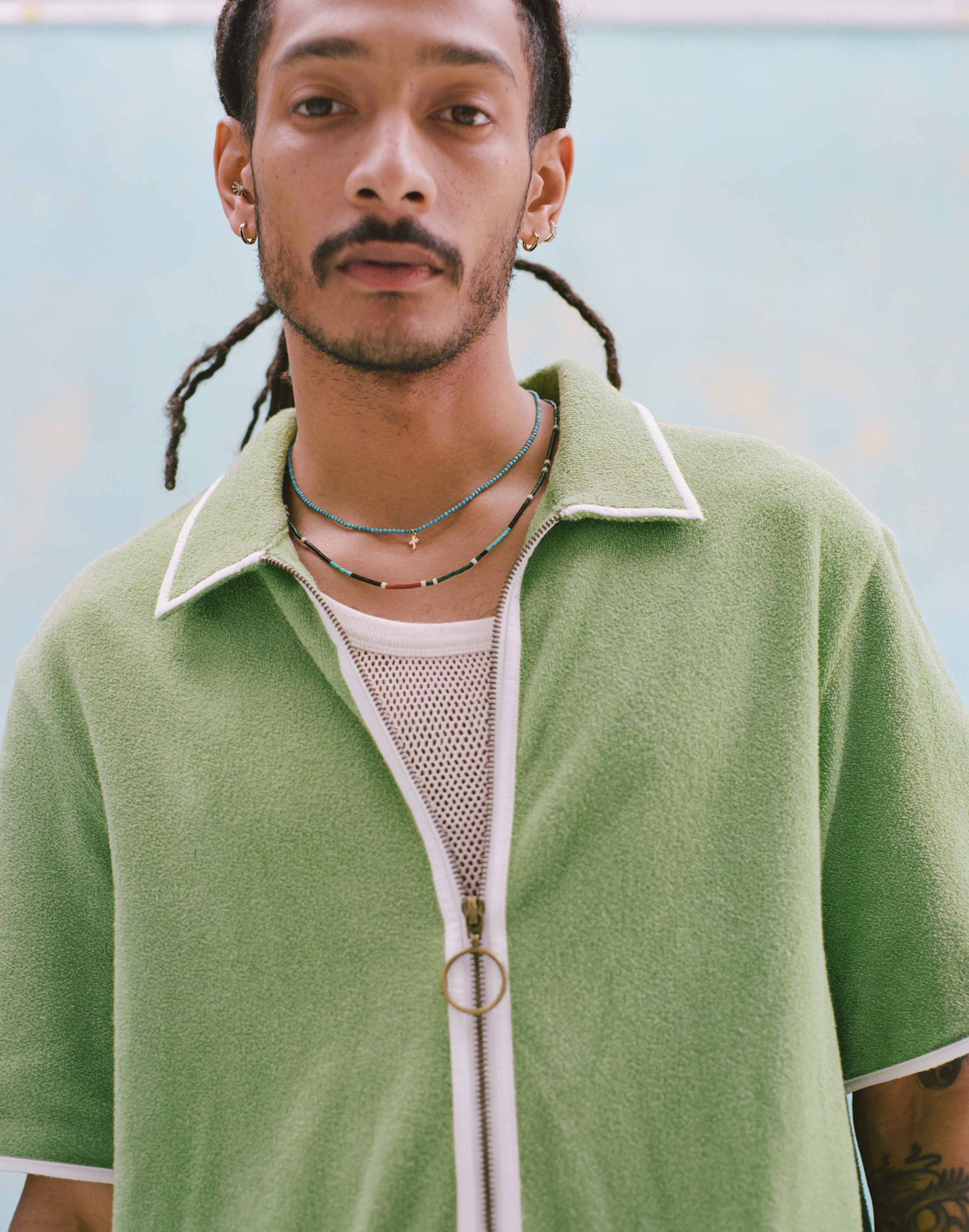 Male model with dreadlocks wearing the 'Standard Issue' Cabana (green) half-zipped with a mesh, white tank top beneath it, and two beaded multicolored necklaces around his neck, shot on a light blue background.